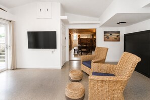 A modern living room with a flat-screen TV, wicker furniture, and a dining area in the background.