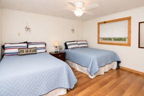 A bedroom with two beds, a ceiling fan, a bedside table with a lamp, and a window with blinds.