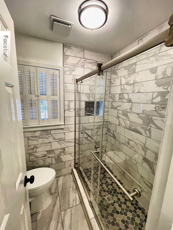 Un baño con cabina de ducha de vidrio, inodoro y ventana con persianas.