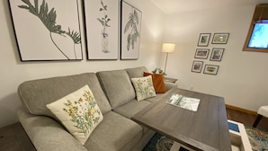 A modern living room with a grey sofa, decorative pillows, a wooden coffee table, and framed botanical prints on the wall.