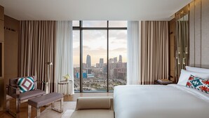 A modern hotel room with a large bed, a bench, a small table, and a view of a cityscape through floor-to-ceiling windows. - The Ritz-Carlton, Baku (Baku)