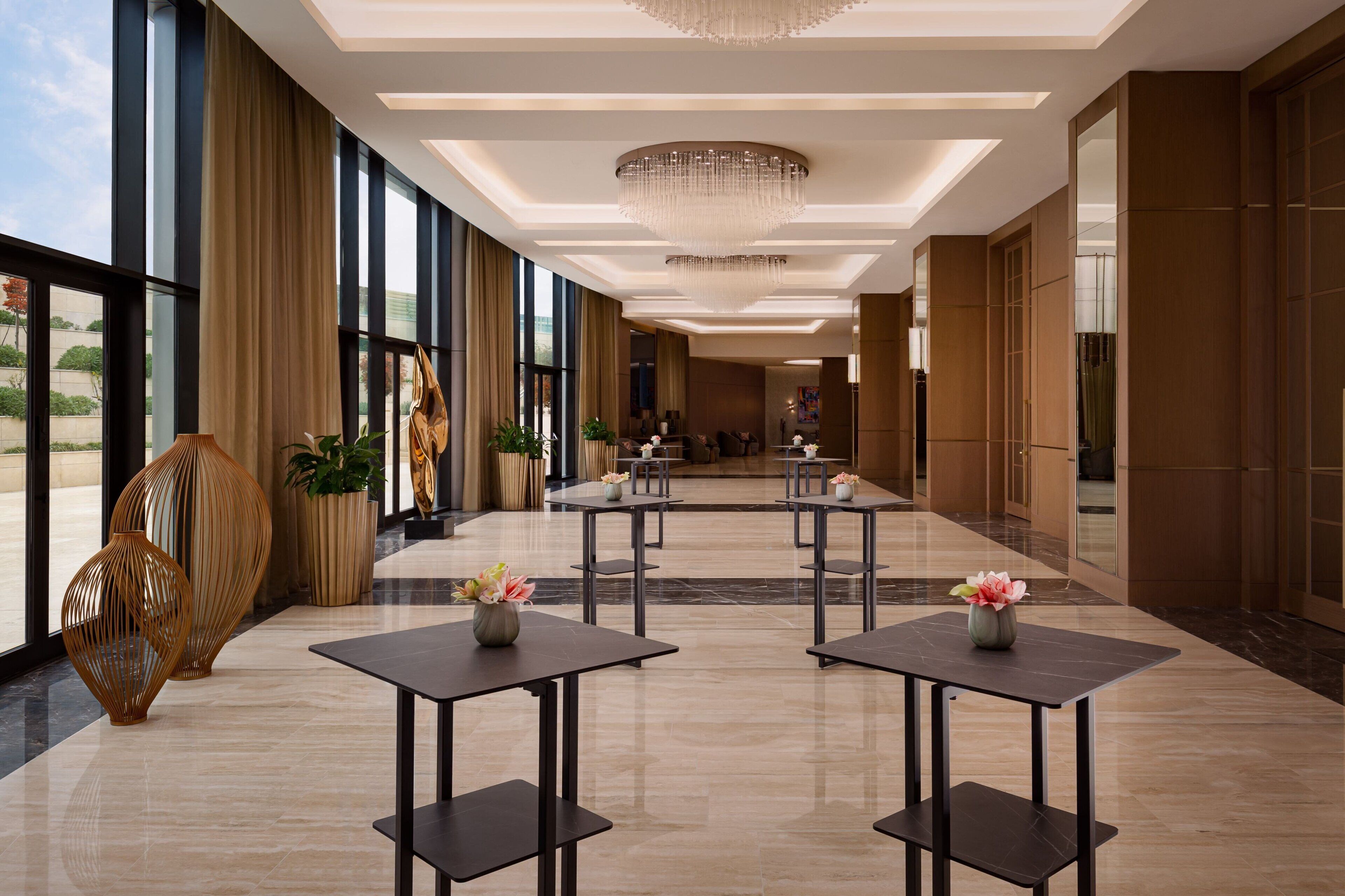 Um hall de hotel espaçoso, com pé direito alto, grandes janelas e um candelabro. Mesas e cadeiras dispostas para os hóspedes.