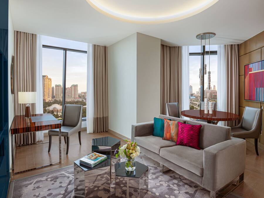 Quarto de hotel moderno com uma janela grande oferecendo vista para a cidade, um sofá com almofadas coloridas, uma mesa de jantar com cadeiras e uma TV de tela plana.