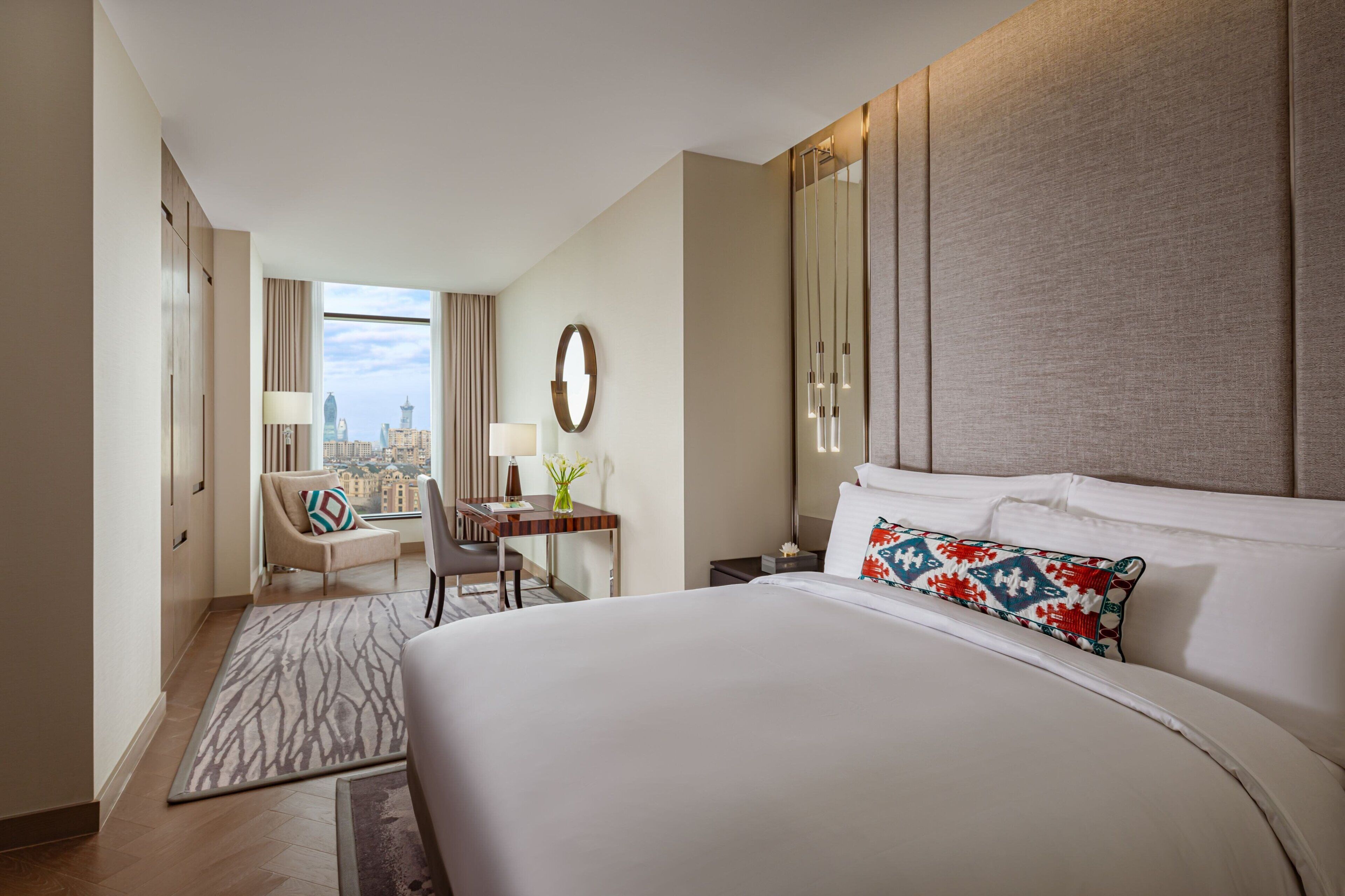 A modern hotel room with a large bed, a desk, a chair, and a view of the cityscape.