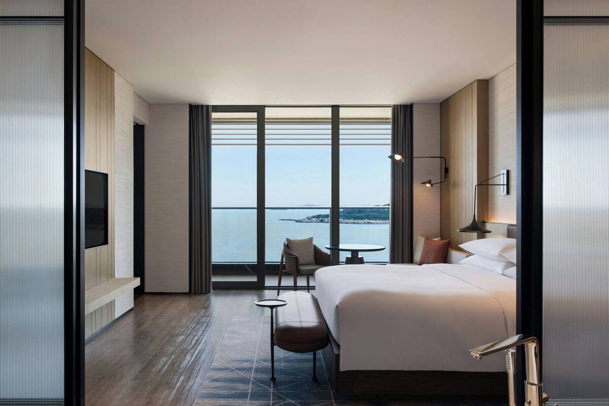 Ein modernes Hotelzimmer mit einem großen Bett, einer Sitzecke mit Tisch und Meerblick durch raumhohe Fenster.