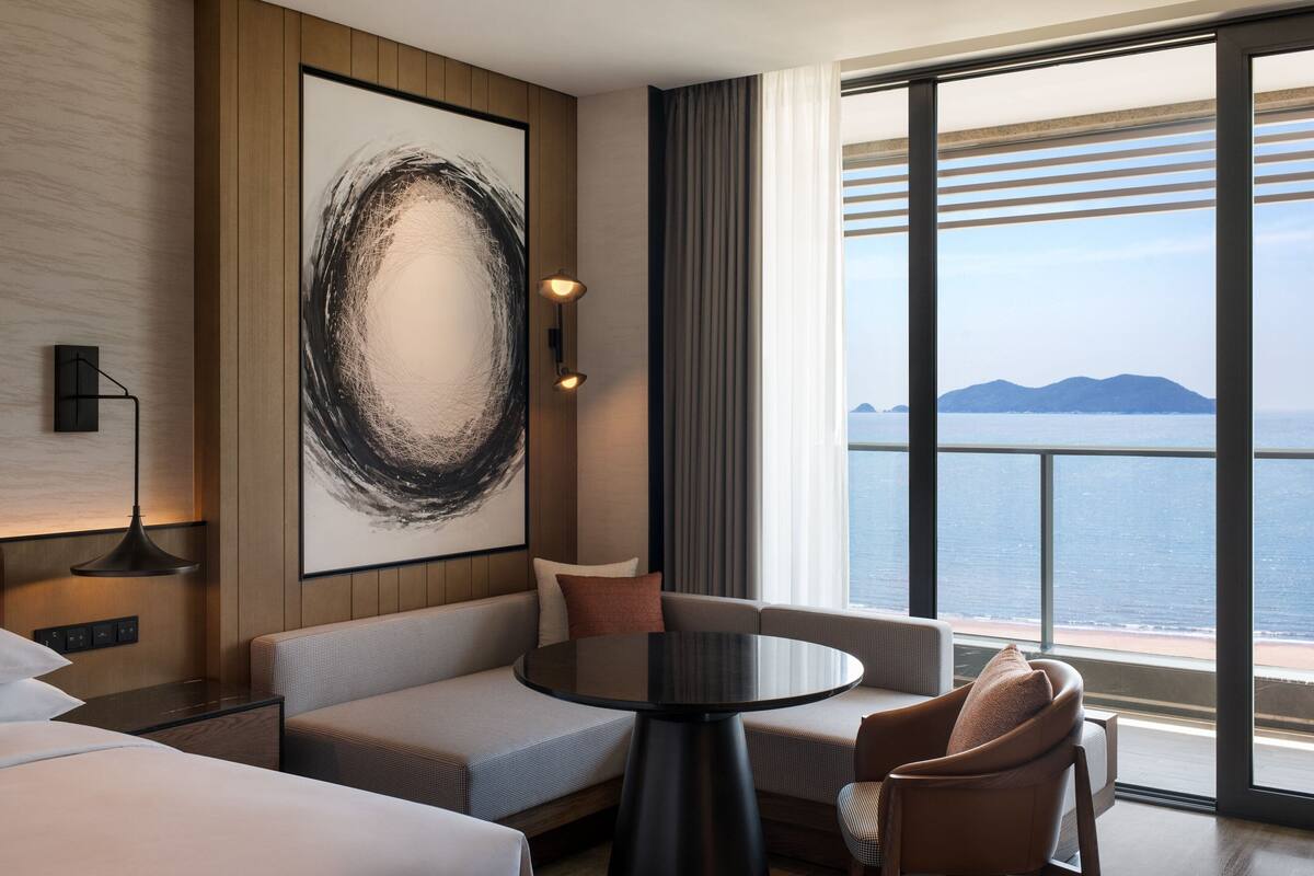 Ein modernes Hotelzimmer mit großem Fenster, rundem Tisch und einer Couch.