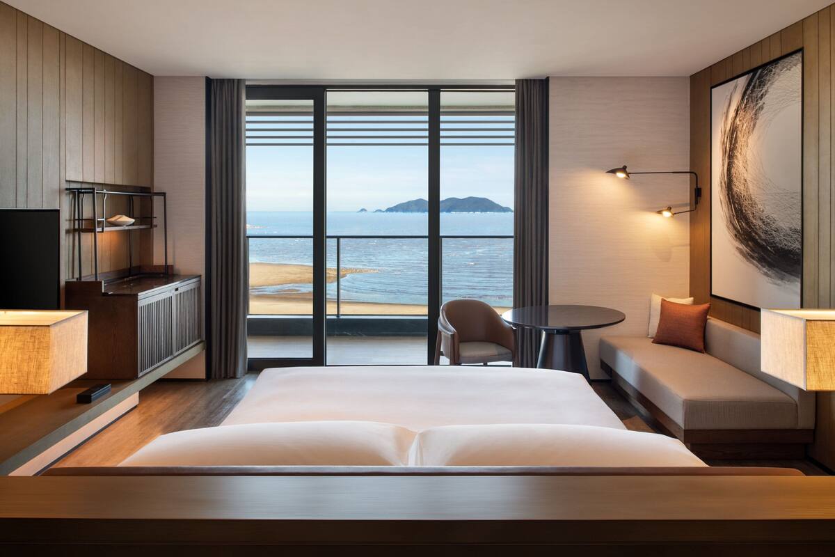 Ein Hotelzimmer mit einem großen Bett, einem Schreibtisch, einem Sessel, einem kleinen Tisch und Meerblick.