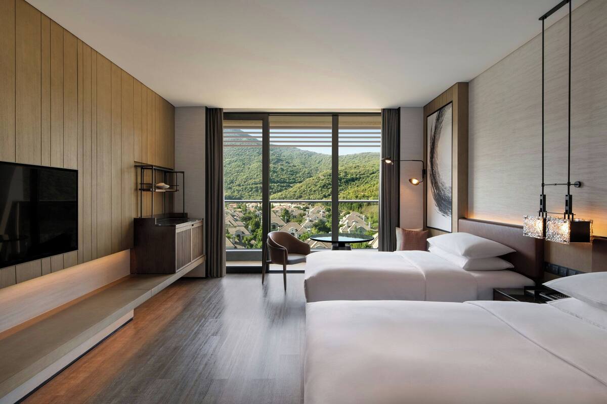 Ein modernes Hotelzimmer mit einem großen Bett, einem Fernseher, einem Schreibtisch und einem Stuhl. Das Zimmer hat ein großes Fenster mit Blick auf eine Berglandschaft.