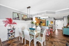 Un comedor con una mesa de vidrio y sillas de mimbre, una cocina al fondo y un pasillo que lleva a una puerta.