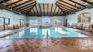 Piscina interior con suelo de baldosas, sillas de descanso y grandes ventanales con vistas.