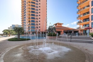 Un hotel con una fuente central, piscina y zona de playa con sillas de descanso.