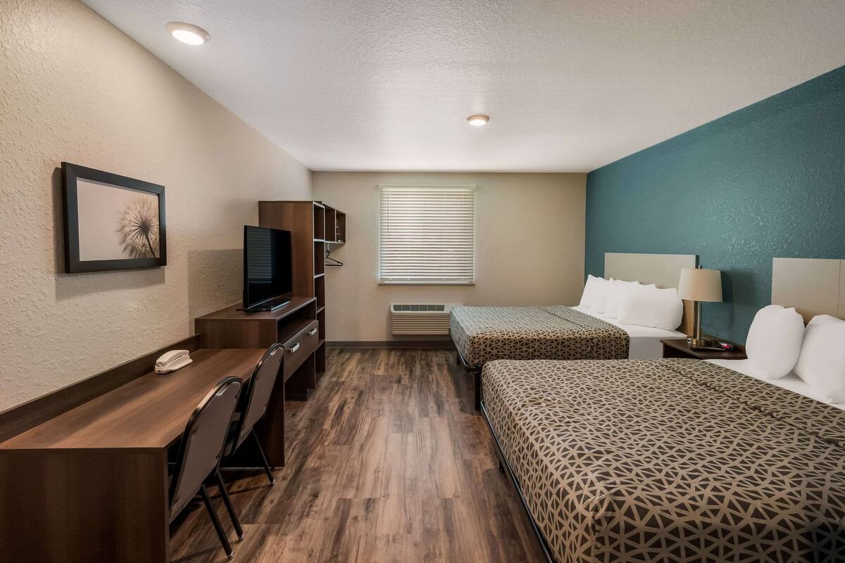 Habitación de hotel con un escritorio de madera, un televisor, una cama con ropa de cama estampada, una mesita de noche con lámpara y una ventana con persianas.