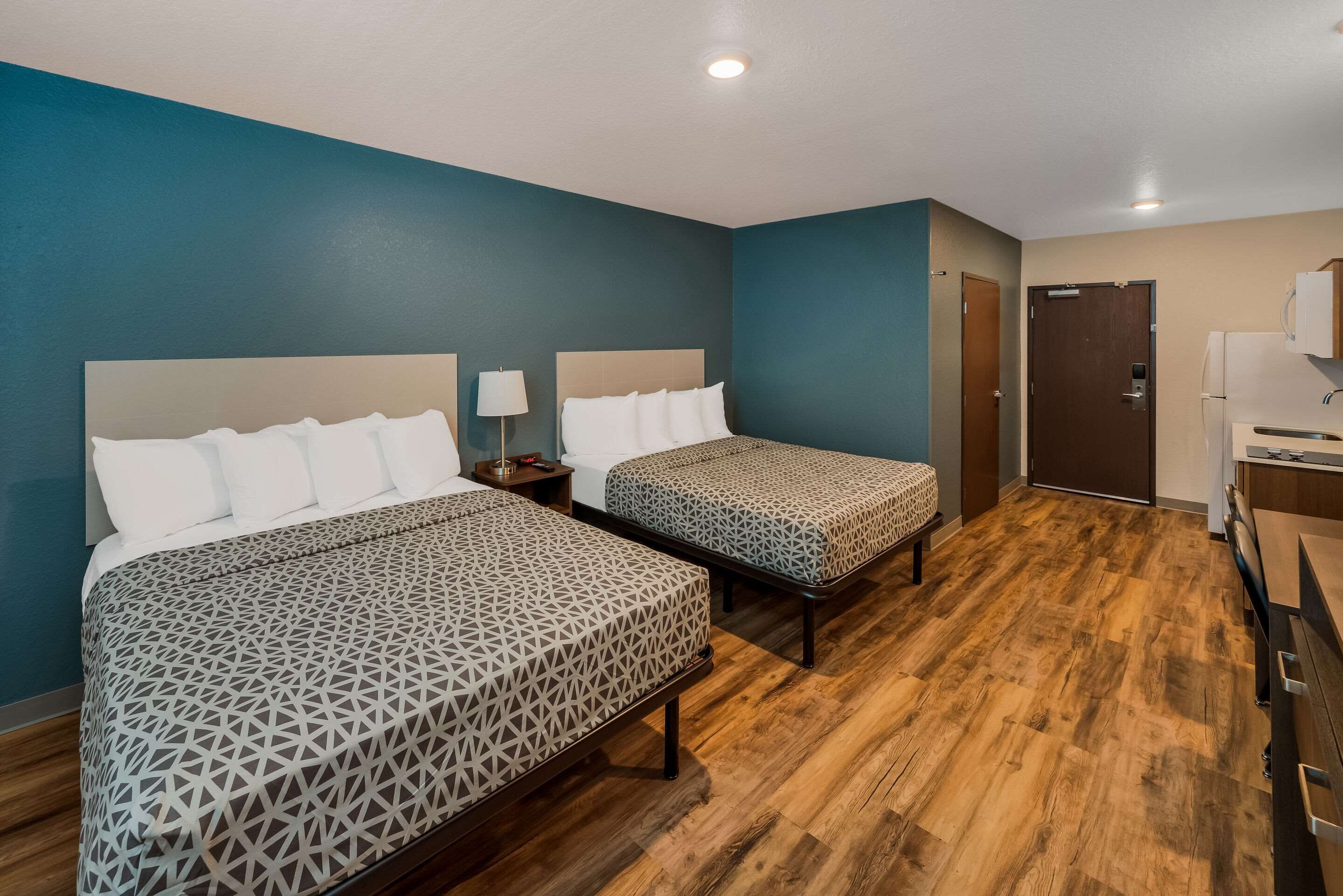 A hotel room with two beds, a wooden floor, a door, and a kitchenette.
