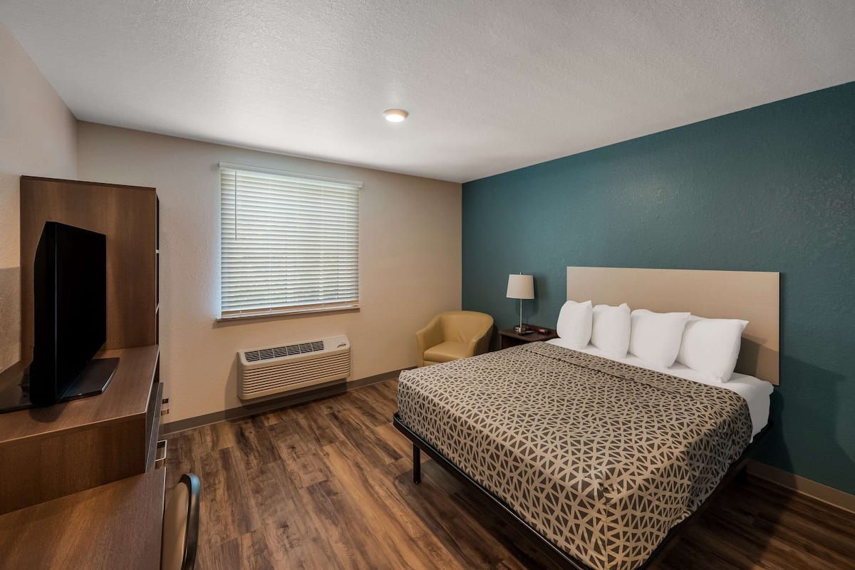 Una habitación de hotel con cama, televisión, silla, lámpara y ventana con persianas.