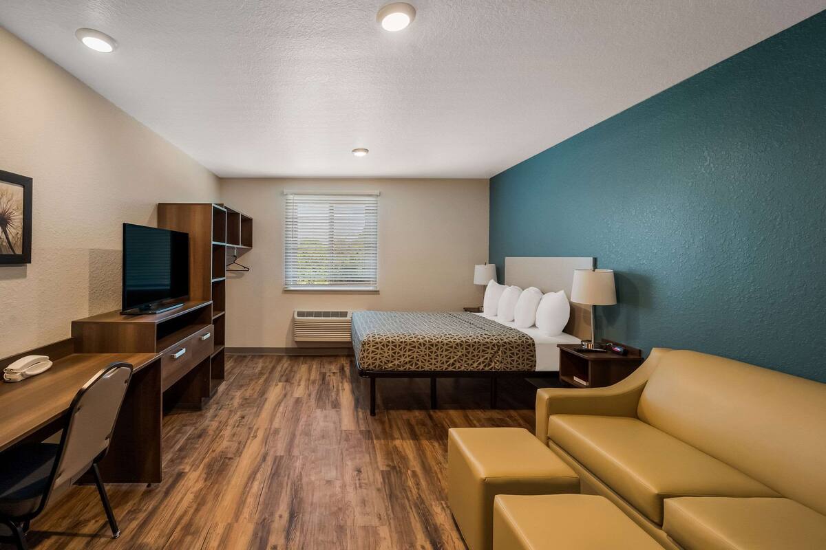Habitación de hotel con cama, escritorio con computadora, televisor, sofá y ventana con persianas.