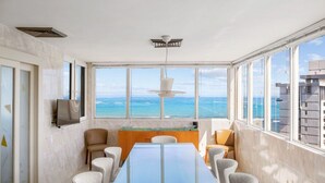 A modern dining area with a large table, chairs, a TV, and a view of the ocean.
