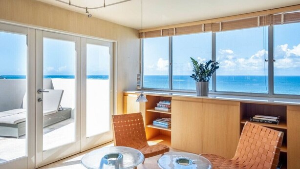 A room with a view of the ocean, a balcony with a chair, and a bookshelf.