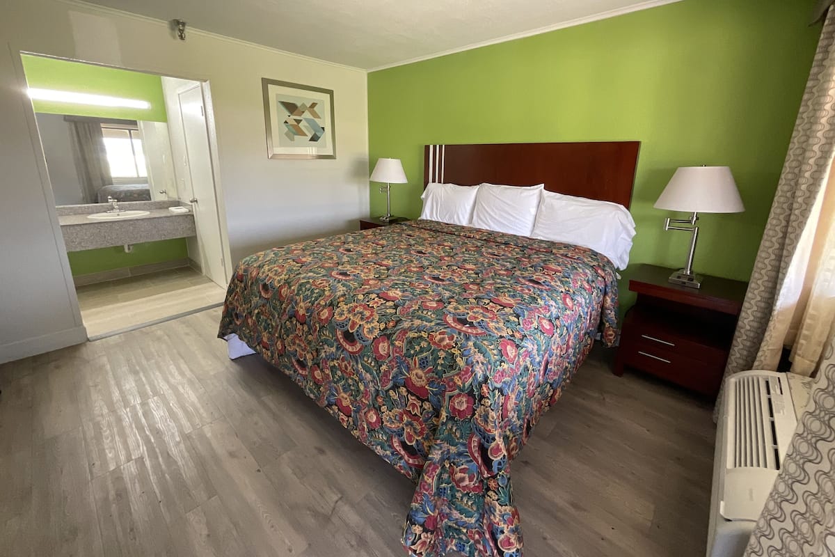 A hotel room with a bed, two bedside lamps, a nightstand, a mirror, a bathroom with a sink, and a green wall.