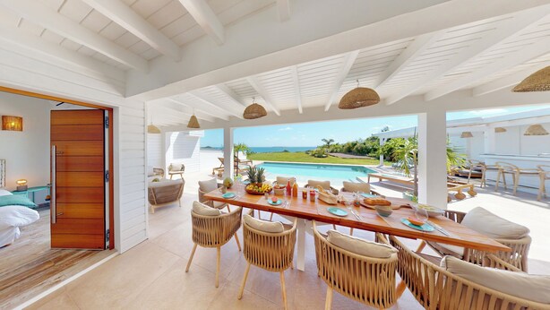 Un comedor amplio con vistas a una piscina y al océano, con mobiliario de mimbre y una mesa de madera lista para comer.