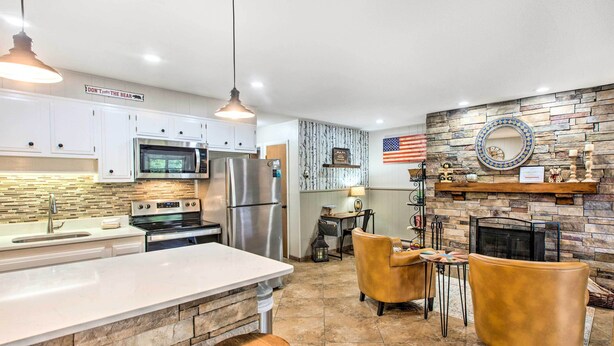 A modern kitchen with white cabinets, stainless steel appliances, and a stone backsplash. There is a dining area with two leather chairs and a small table. An American flag and a decorative mirror are on the wall. - Main St Red River Ski Condo w/ Mountain View! (Red River)