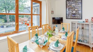 Zona pranzo con tavolo apparecchiato per quattro persone, televisione a parete e vista sul giardino attraverso ampie finestre.