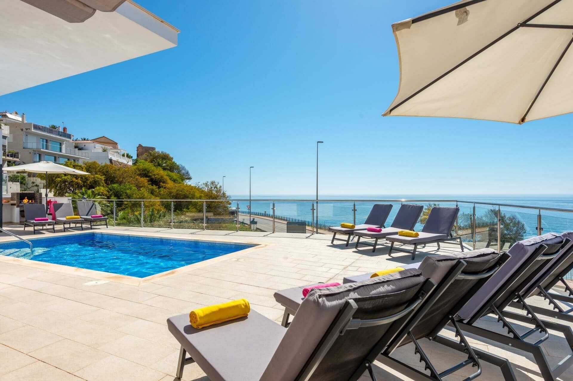 A poolside area with lounge chairs and umbrellas, overlooking a clear blue sea and a coastal town.