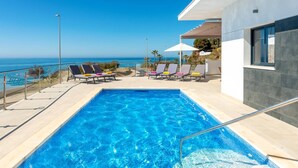 Une piscine rectangulaire à l’eau d’un bleu cristallin, entourée de chaises longues avec des coussins, d’une pergola blanche et offrant une vue sur l’océan.