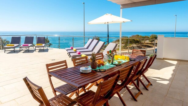 Une terrasse avec des tables et des chaises en bois, un parasol blanc et une vue sur l’océan.