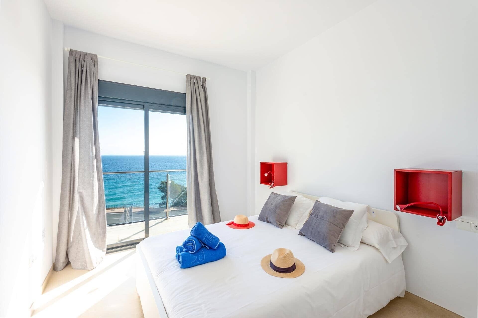 A hotel room with a large window, a bed with white linens, two red wall sconces, and a view of the sea.