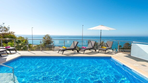 A swimming pool with lounge chairs and an umbrella, overlooking a clear blue sea.