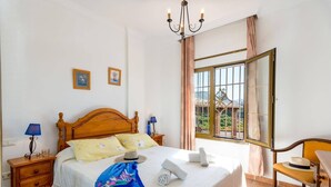Een slaapkamer met een houten bed, twee nachtkastjes, een kroonluchter, een raam met gordijnen en uitzicht op groen.