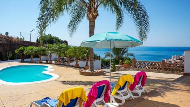 Zona junto a la piscina con sillones de descanso y una sombrilla grande, con vistas a la costa.