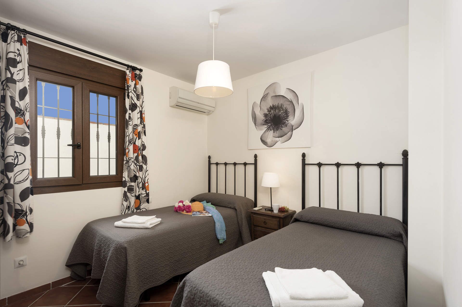 Ein Schlafzimmer mit zwei Betten, einem Fenster mit gemusterten Vorhängen, einer Wand-Klimaanlage, einer Nachttischlampe und einem großen Blumenbild.
