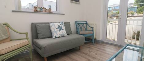 Una habitación con un sofá gris, una silla azul, una mesa de vidrio y una ventana con vistas a edificios.