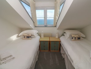 Quarto pequeno e bem iluminado com duas camas, uma mesinha de cabeceira e uma janela com vista para o mar.