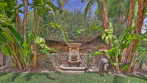 En trädgård med en fontän av sten, omgiven av bananplantor och en mur av tegelsten.