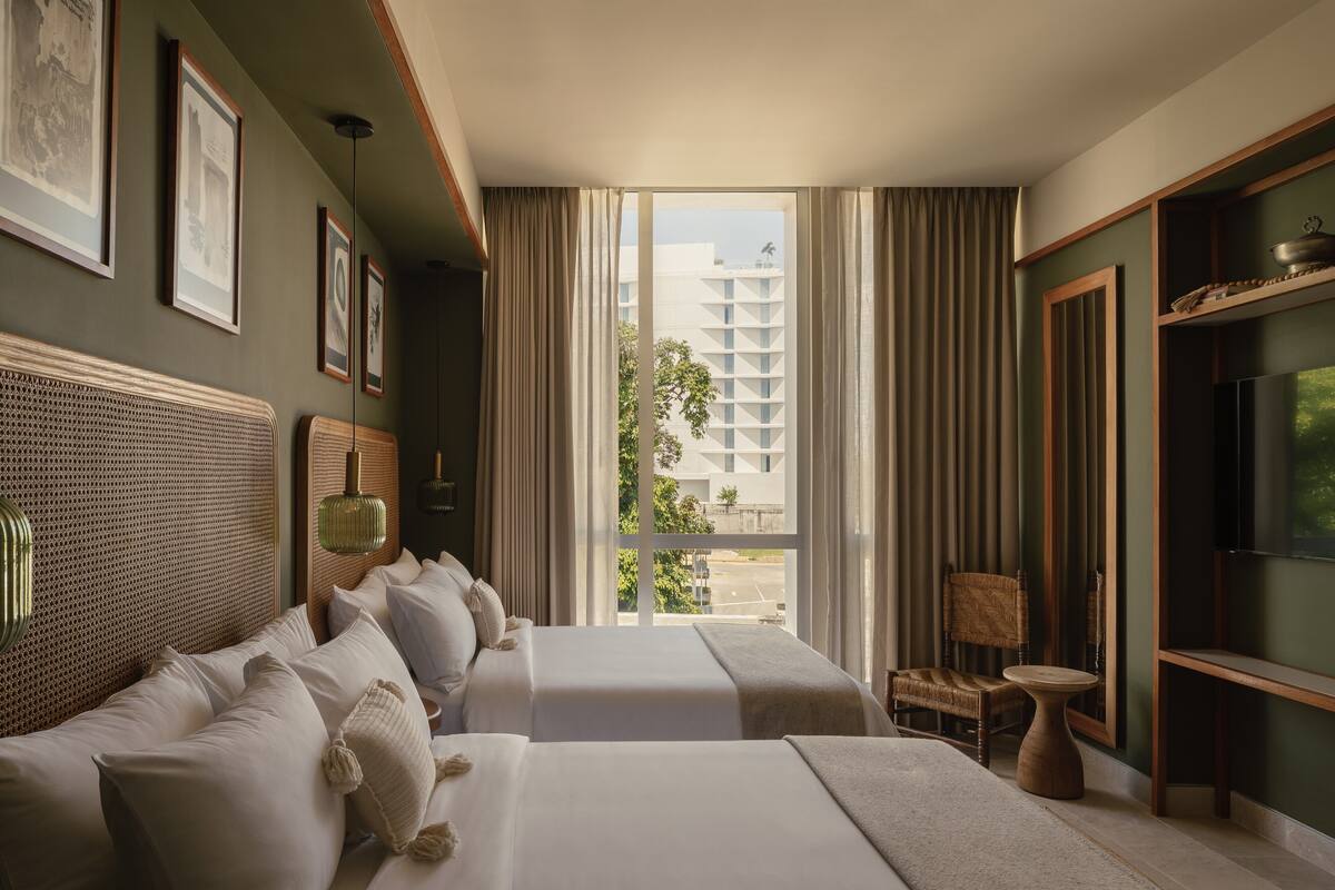A hotel room with two beds, a large window with curtains, a television, and framed pictures on the wall.