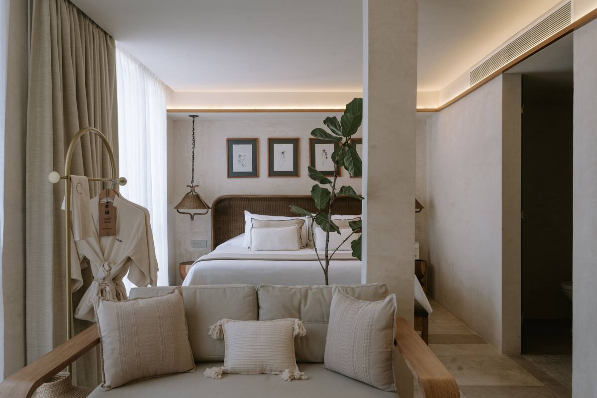 A hotel room with a bed, a sitting area with cushions, a hanging robe, and a large plant.