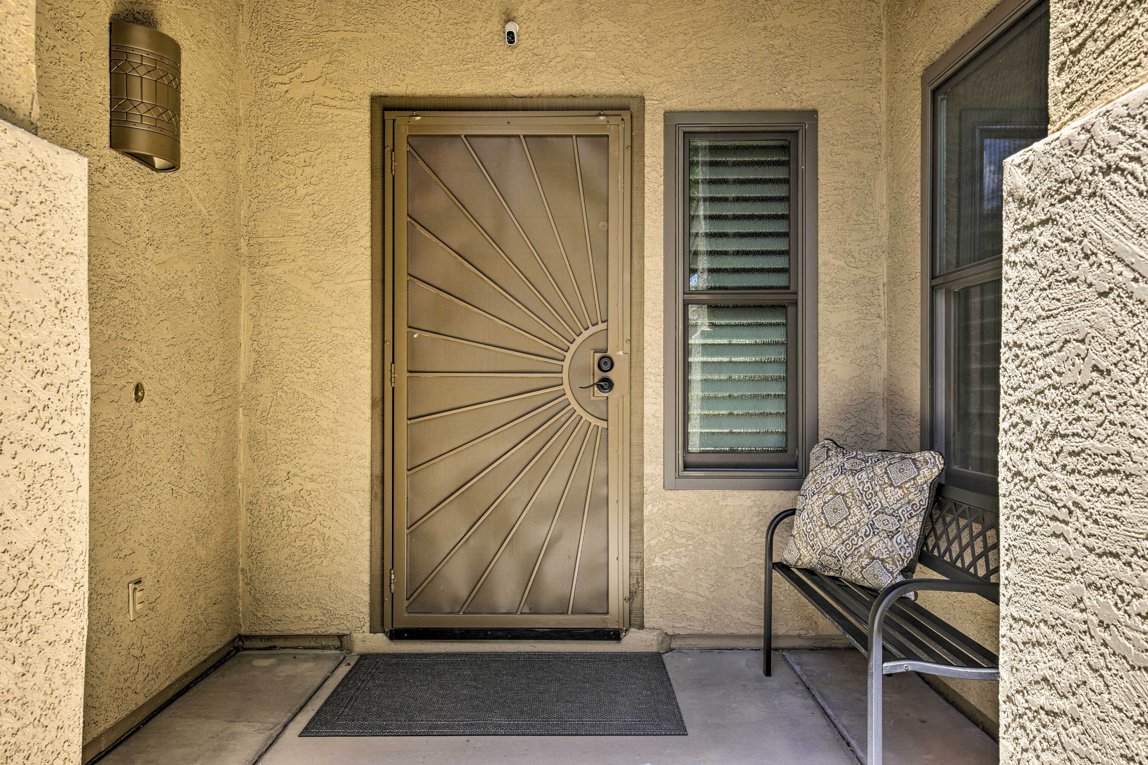Una pared beige con textura, una puerta metálica con diseño de sol, un banco con cojín estampado y una ventana con persianas horizontales.