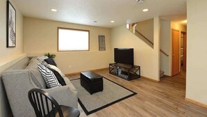 A living room with a sofa, a black ottoman, a TV on a stand, and a window. - Updated Twin Home near Sanford Medical Center (West Fargo)