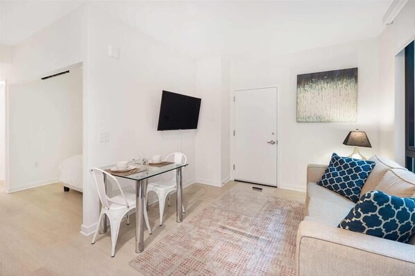 A modern living room with a glass dining table, white chairs, a sofa with patterned cushions, a flat-screen TV mounted on the wall, and a door leading to another room.