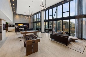 Een ruime woonkamer met een tafelvoetbaltafel, een bank en een salontafel. Er is een keuken met een eettafel en stoelen, en een groot raam met uitzicht op de stad.