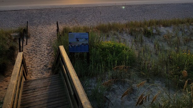 Una pasarela de madera que lleva a una playa con un atardecer al fondo.