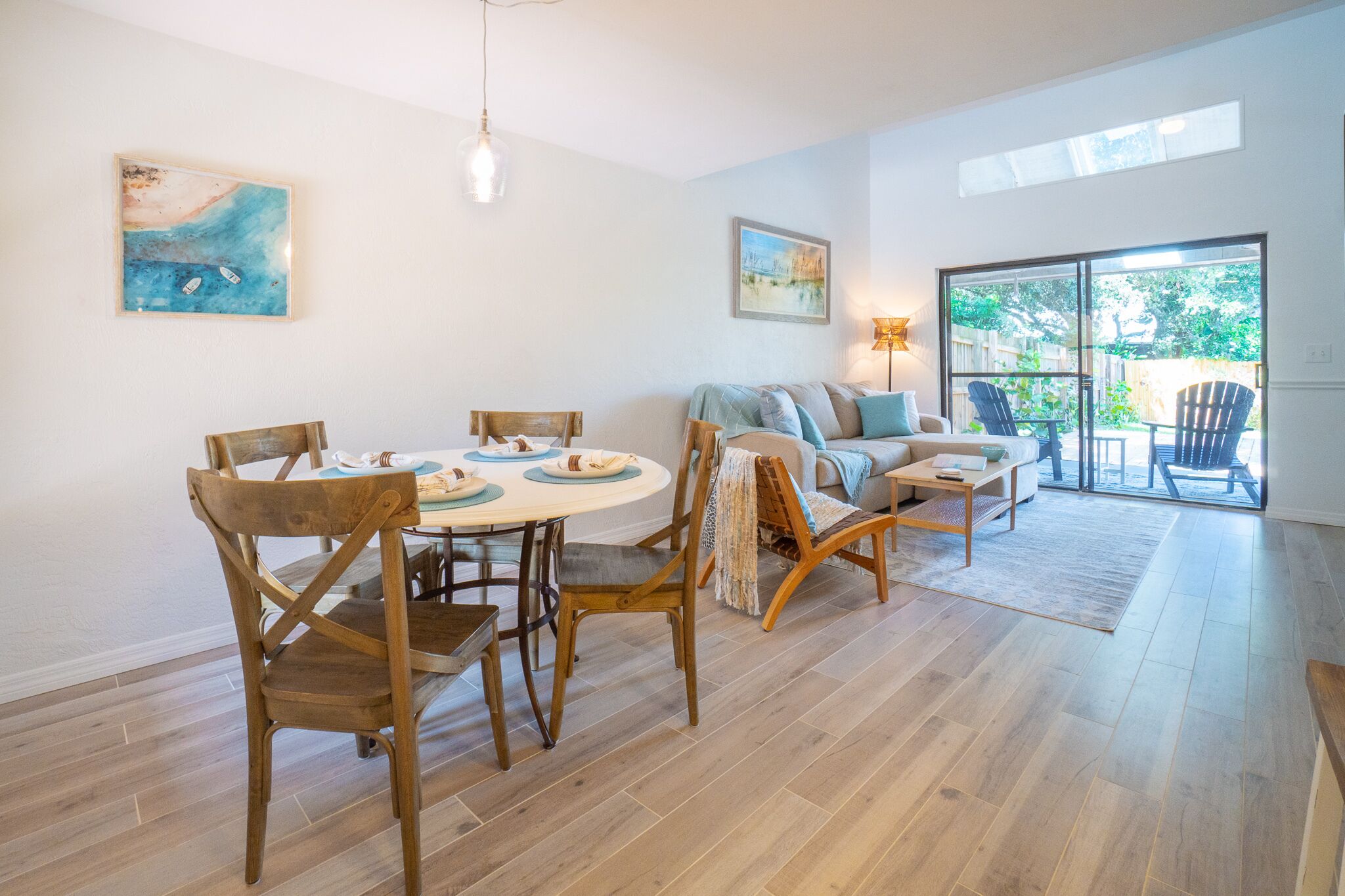 A spacious living area with a dining table, chairs, a sofa, and a coffee table. There are two paintings on the walls and a large window providing a view of the outdoors.