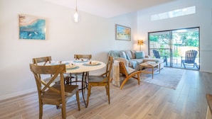 Amplia sala de estar con mesa de comedor, sillas, sofá y una mesa de centro. Hay dos cuadros en las paredes y un ventanal con vistas al exterior.