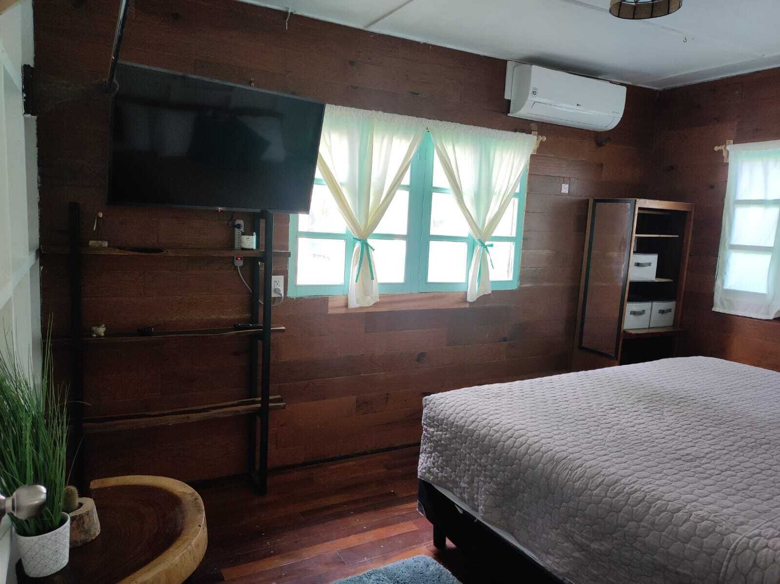 A wooden room with a bed, a TV mounted on the wall, a wardrobe, and a window with curtains.