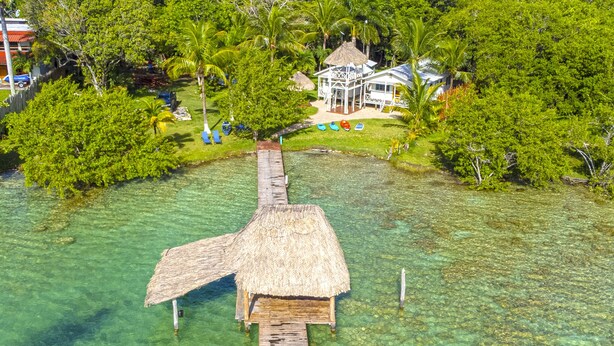 A thatched-roof hut on a wooden pier extending into clear water, surrounded by lush greenery and a white building with a thatched roof in the background. - Beautiful Cabin with private dock in Bacalar Lake (Buenavista)