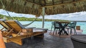 A wooden deck with a thatched roof, a lounge chair, a dining table with chairs, and a view of the sea. - Beautiful Cabin with private dock in Bacalar Lake (Buenavista)