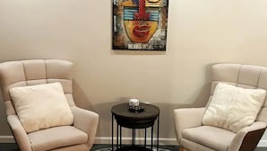Deux fauteuils beiges avec des coussins blancs, une petite table noire ornée d’un objet décoratif et, au mur, un tableau abstrait aux couleurs vives.