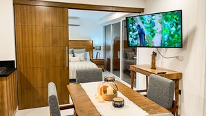 Quarto de hotel moderno com guarda-roupa de madeira, uma mesa de jantar com cadeiras estofadas cinzentas, uma TV na parede e um espaço de estar acolhedor com uma pequena mesa e almofadas.